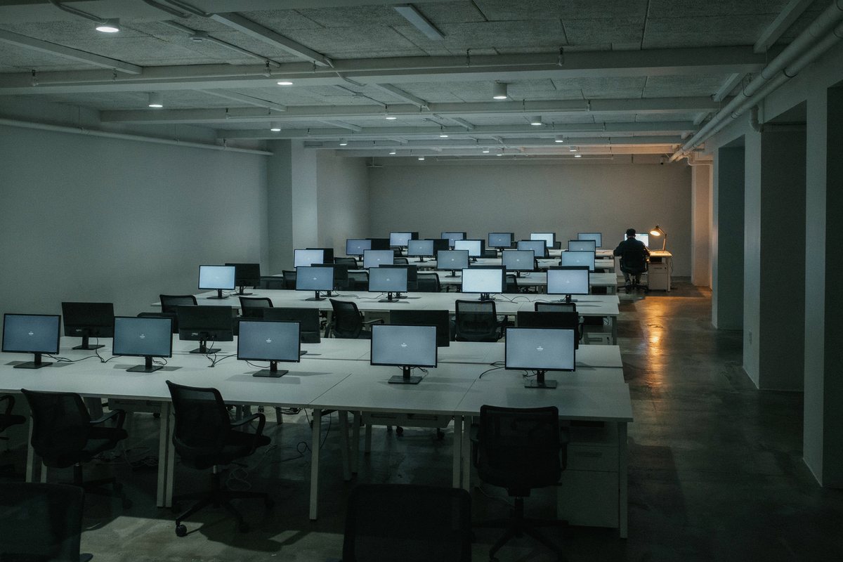 Großraumbüro am Abend, viele Bildschirme zeigen den Anmeldebildschirm, nur eine Person arbeitet noch