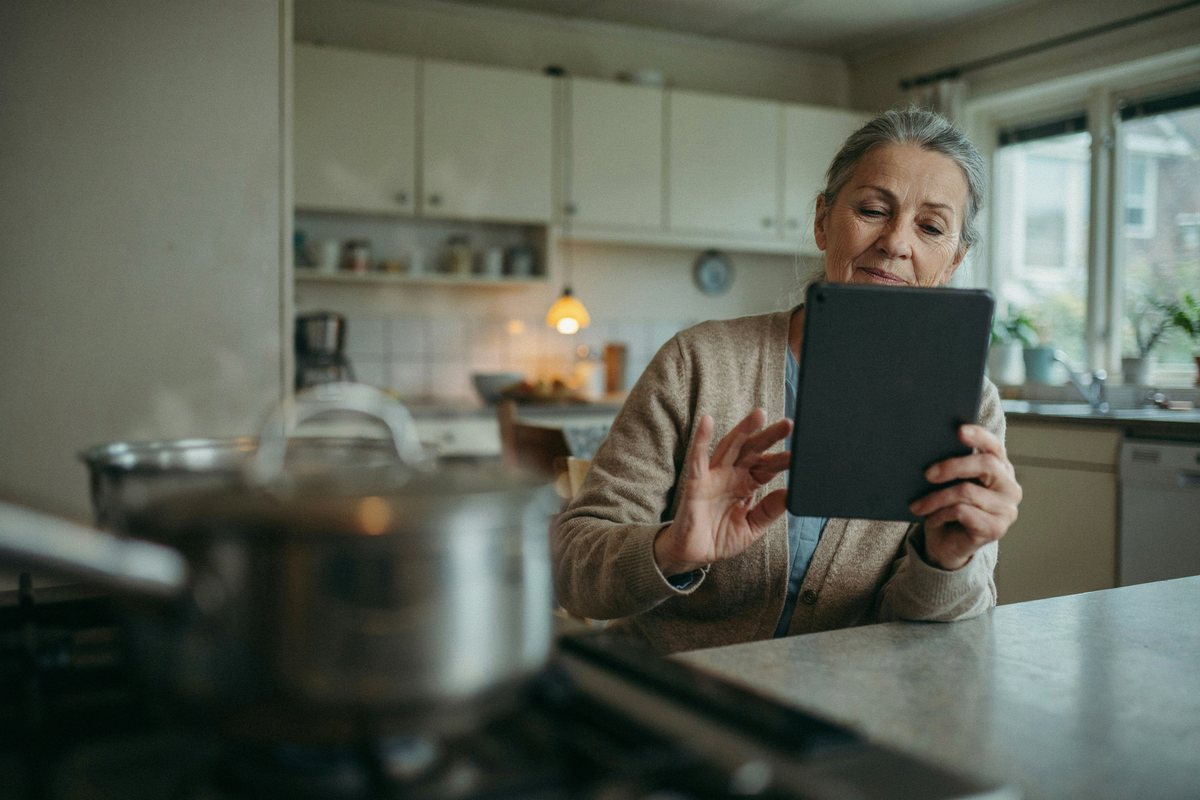 Ältere Frau am Küchentisch spricht mit einem Tablet, im Hintergrund ein Topf auf dem Herd