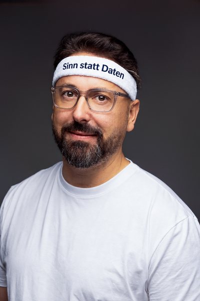 Niko Martzy mit Stirnband „Sinn statt Daten“ im weißen T-Shirt, Porträt vor grauem Hintergrund